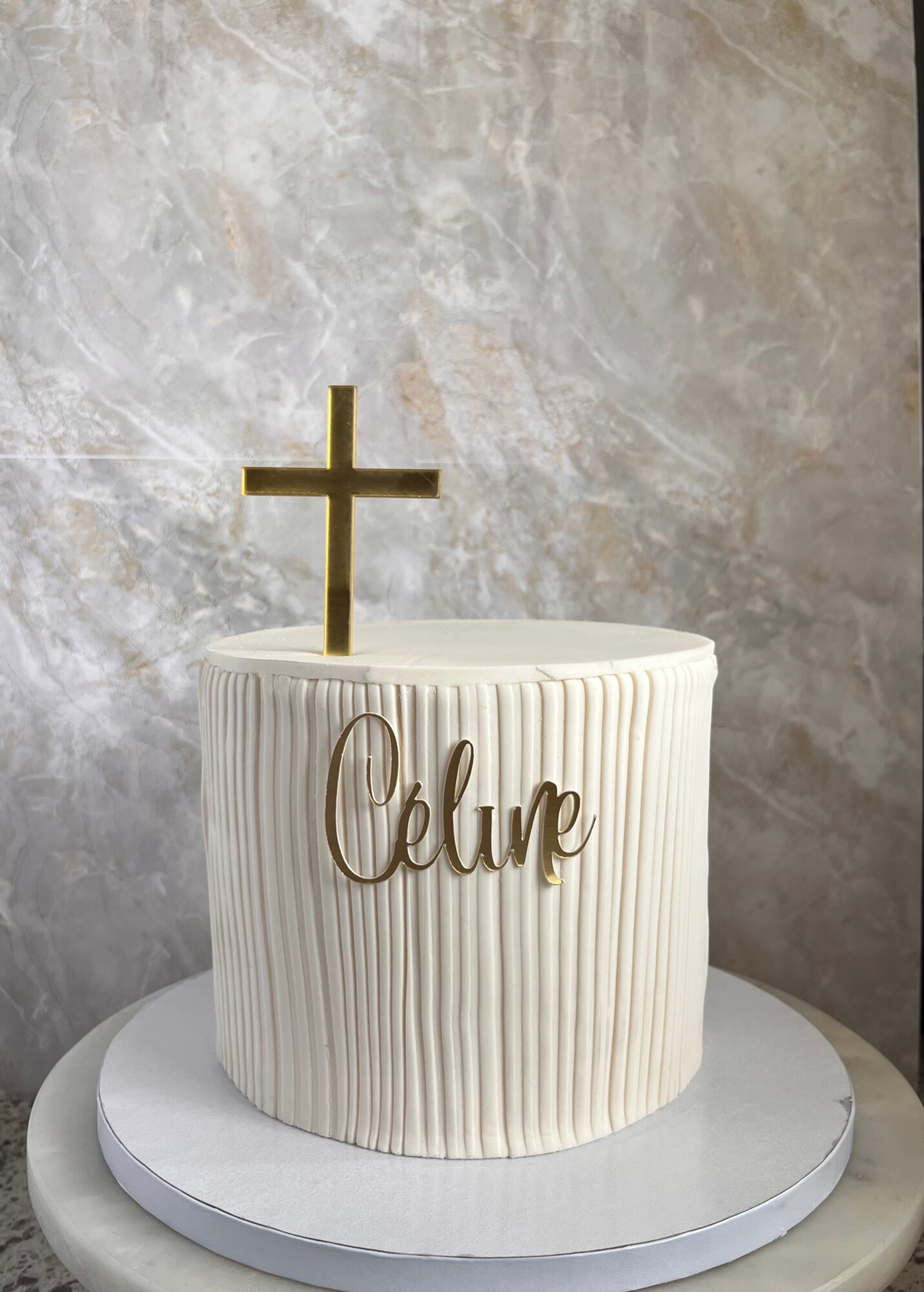 A white cake with vertical ridges, a gold cross on top, and the name 'Oclue' in gold script, on a white base.