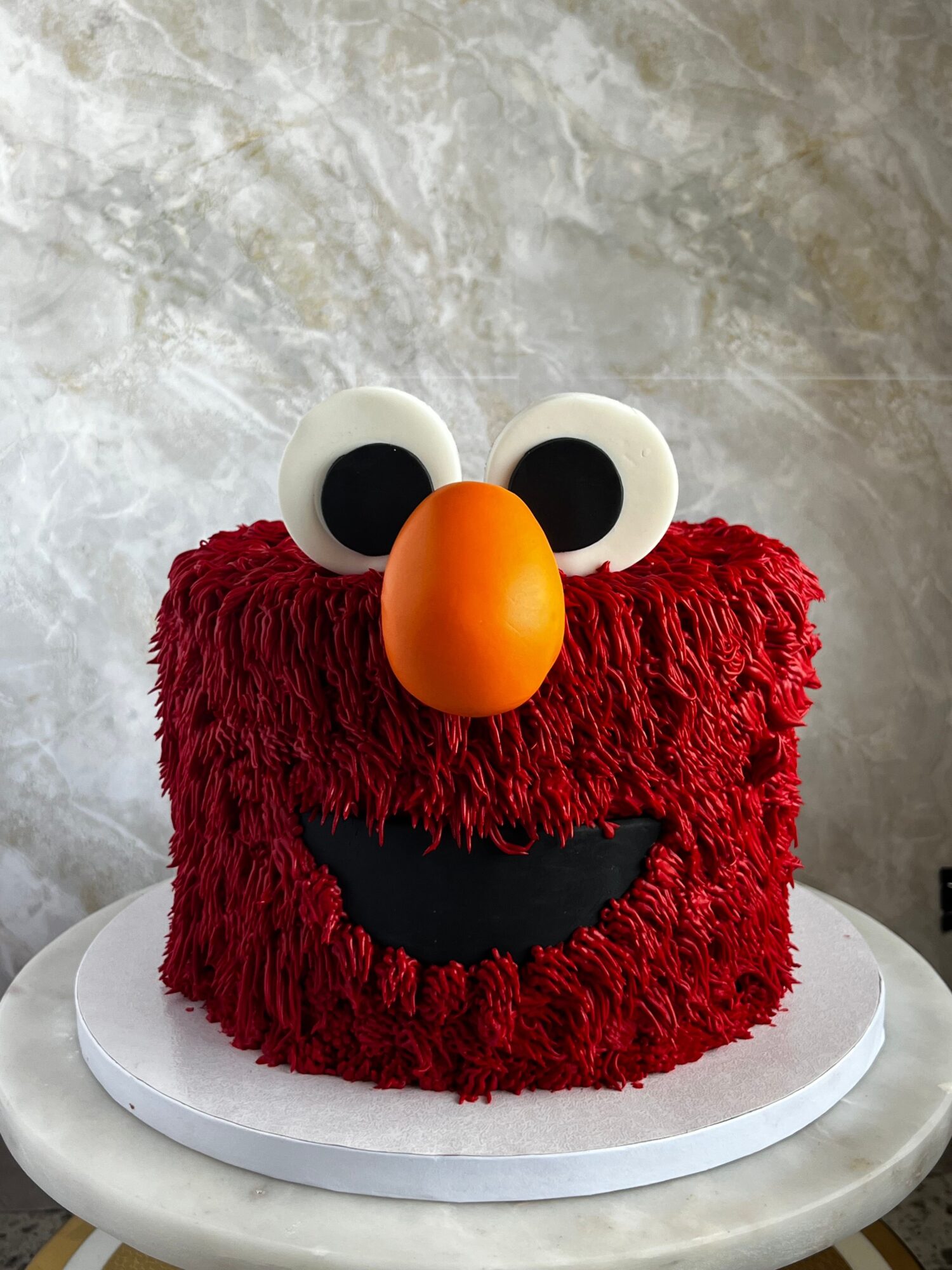 Cake resembling Elmo with red fur, large eyes, orange nose, and a smiling mouth, on a white surface.