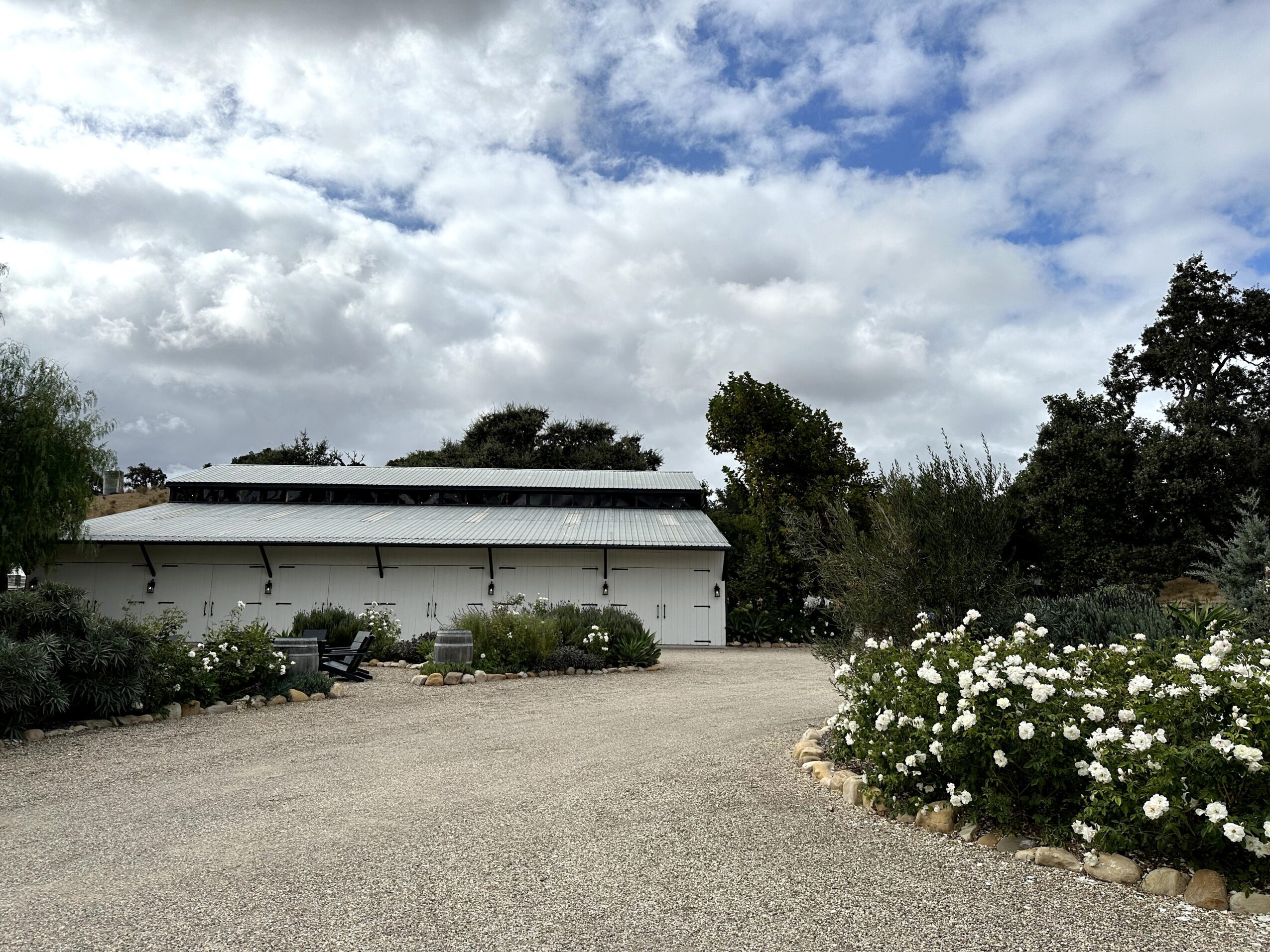 Building with a white exterior, surrounded by trees and flowering bushes, under a partly cloudy sky.