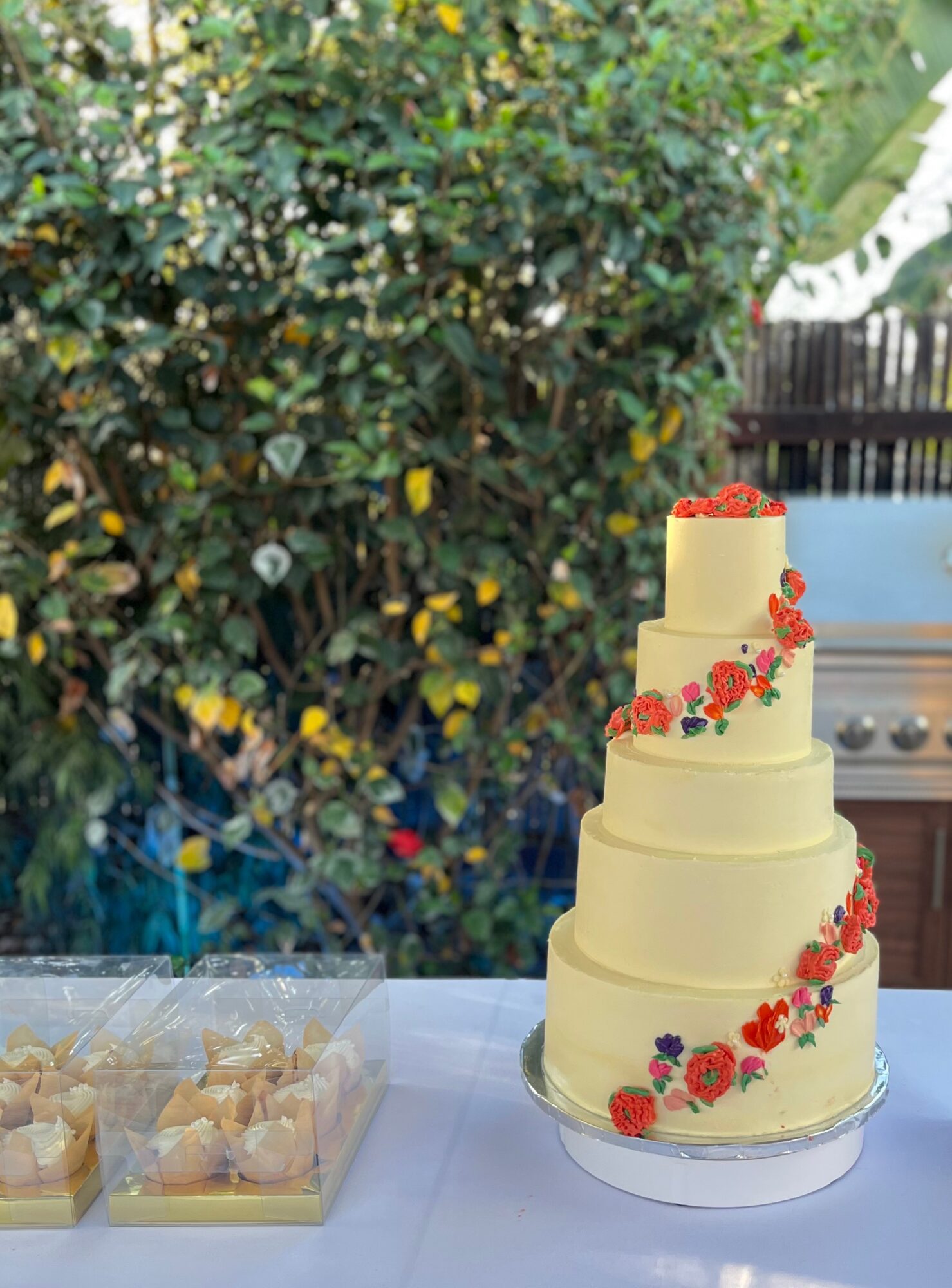 Three-tiered cream-colored cake decorated with red and purple flowers, topped with red berries, on a table outdoors.