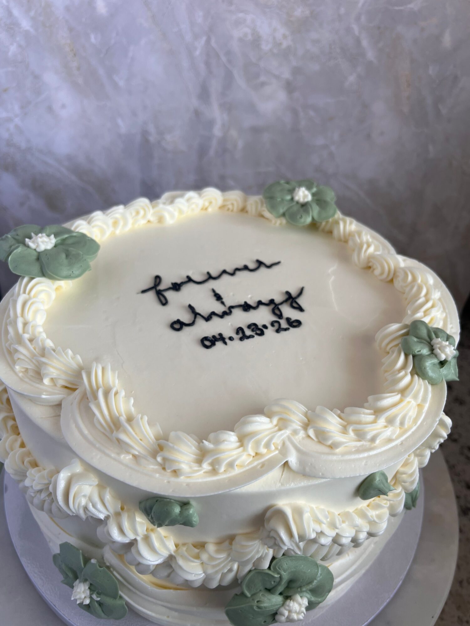 Round cake with white frosting and decorative green flowers, with handwritten message and date on top.