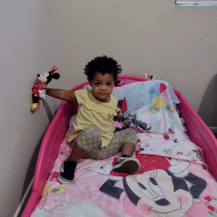 Child sitting on a bed with Minnie Mouse bedding, holding a Minnie Mouse plush toy, smiling at the camera.