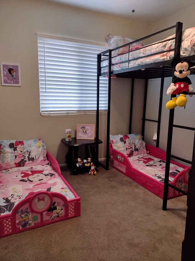 Children's bedroom with two beds, a bunk bed and a bed with pink and floral bedding, a window with blinds, and a small table.