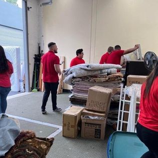People in red shirts working with stacked boxes and blankets inside a warehouse or storage area.