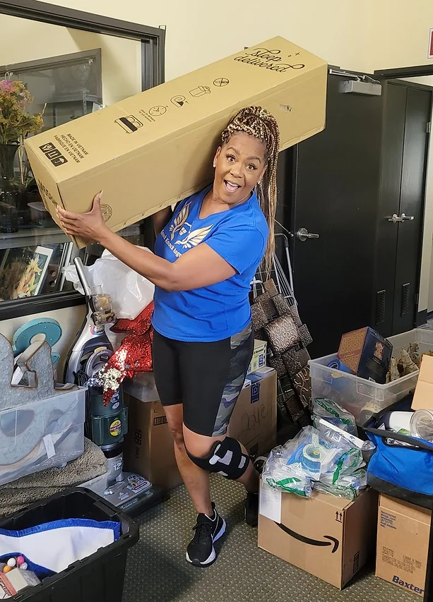 Girl in blue shirt holding a long box, surrounded by boxes and items in a cluttered room.