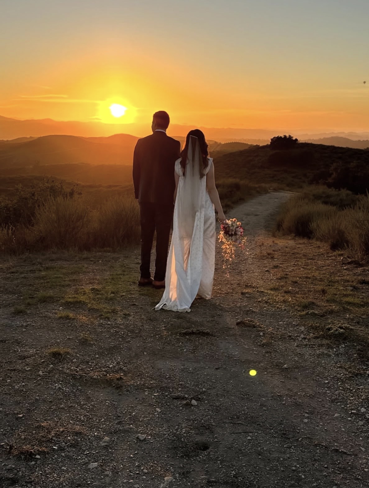 Silhouette of a couple walking on a dirt path during sunset, with a landscape of hills and a colorful sky in the background.