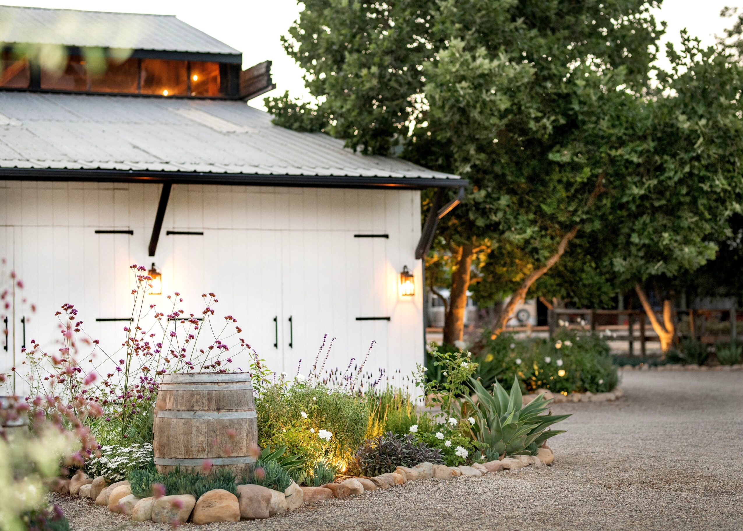 White shed with flowers and a barrel in front, surrounded by trees and a gravel path.