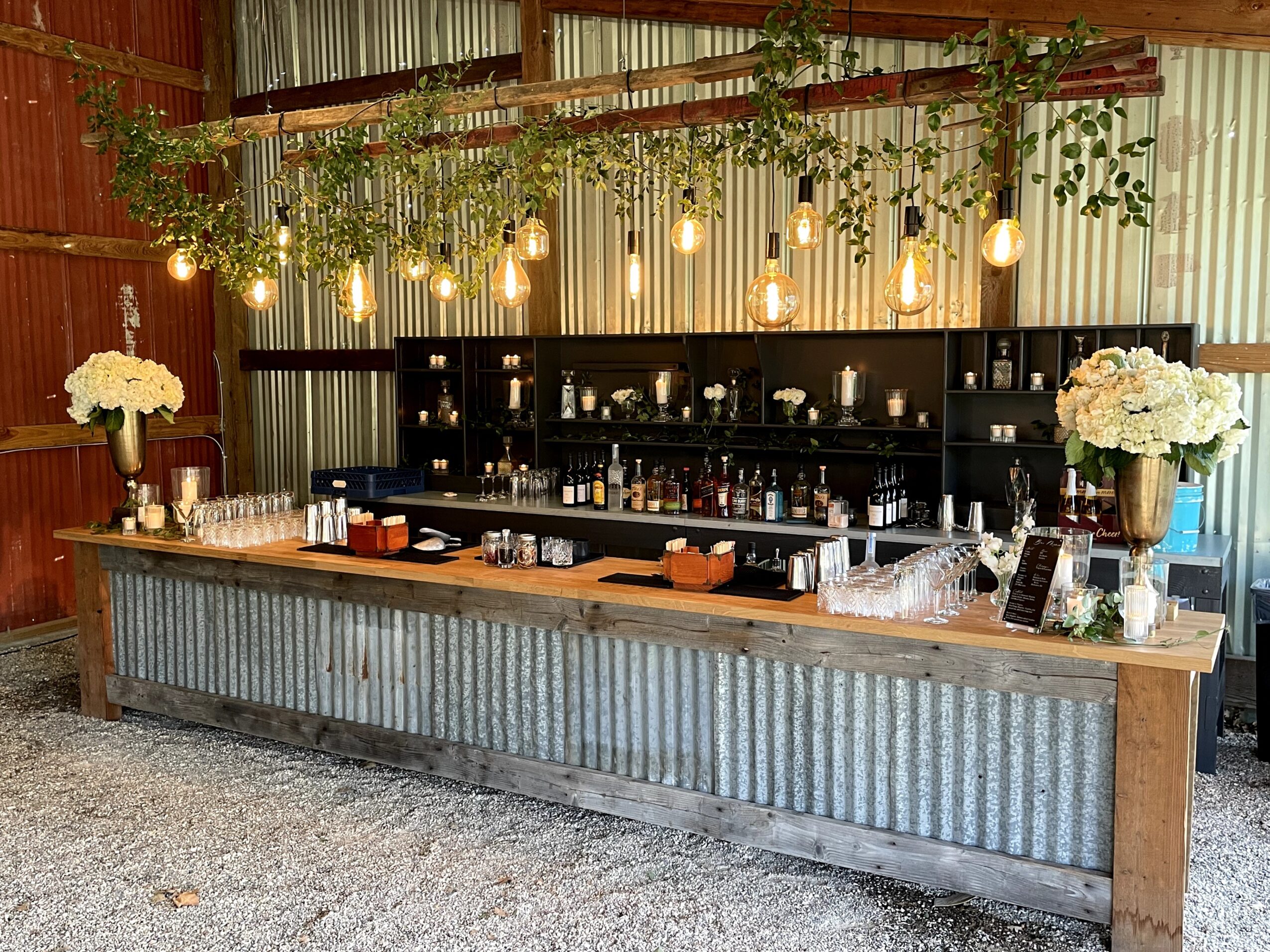 Bar setup with hanging lights, floral arrangements, and a corrugated metal counter in a rustic setting.