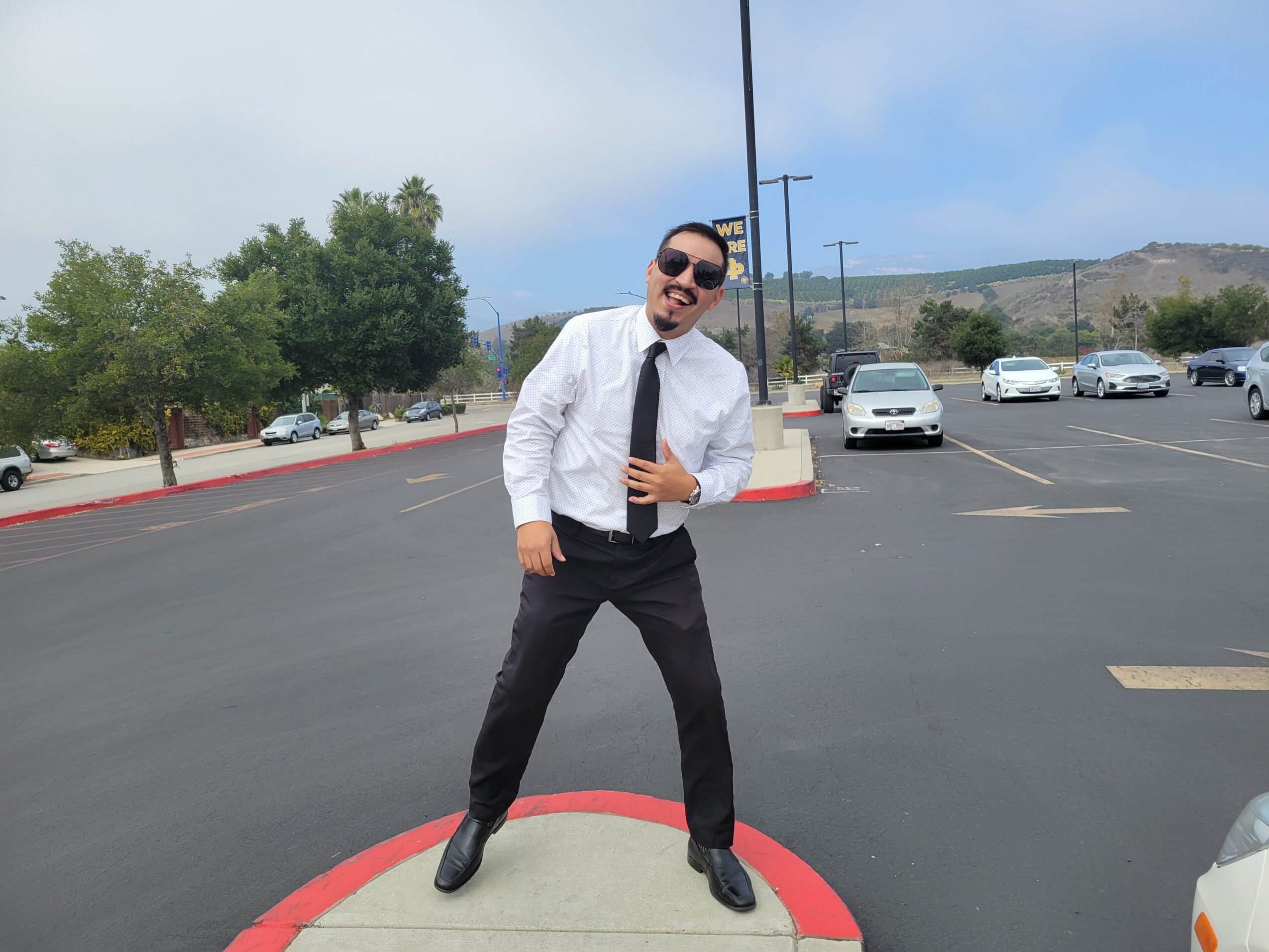 Person in white shirt, black tie, and sunglasses standing on a parking lot curb, smiling, with cars and trees in background.