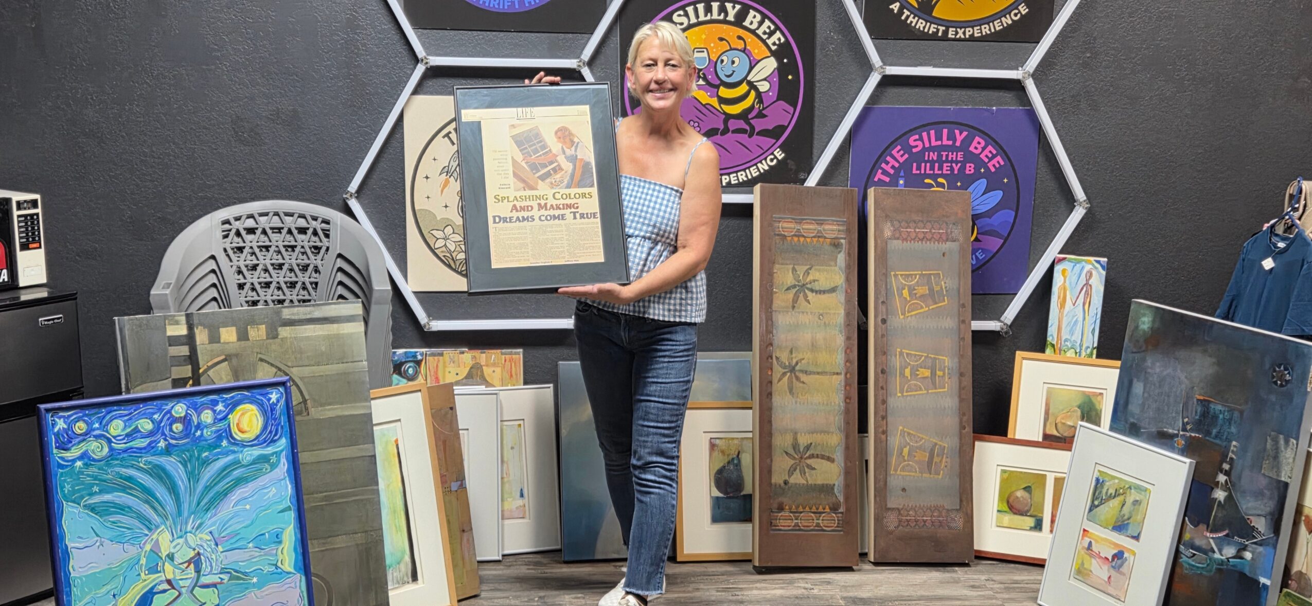 Woman holding framed artwork in an art display room with paintings and decorative items around her.