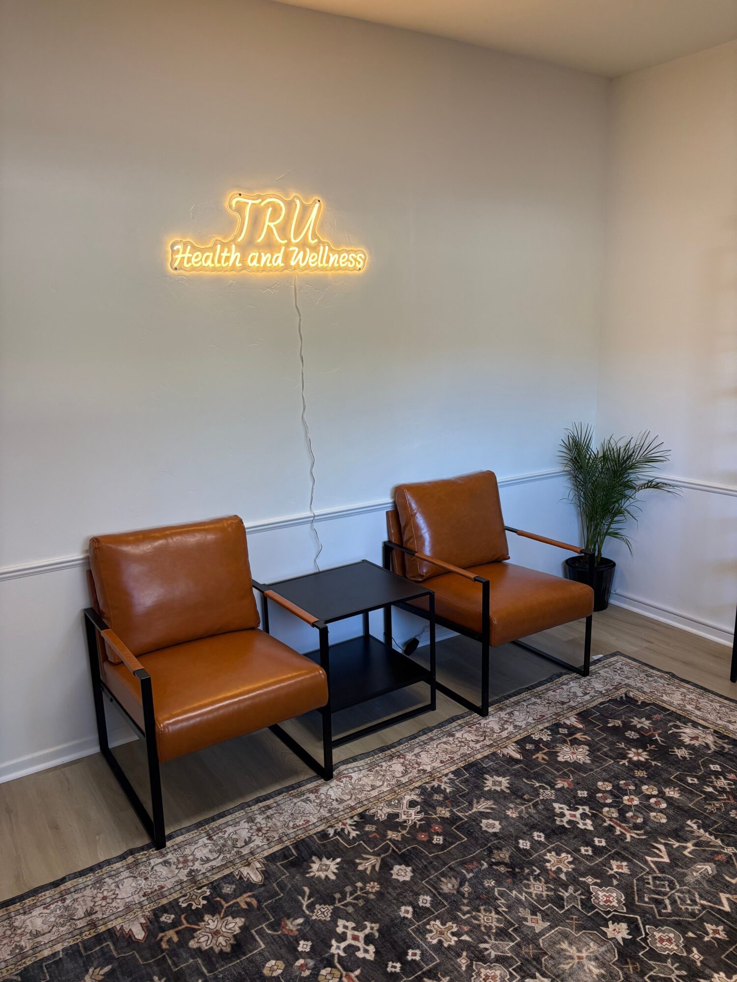 Two brown chairs with black metal frames and a small black table between them, a neon sign on the wall, and a potted plant.