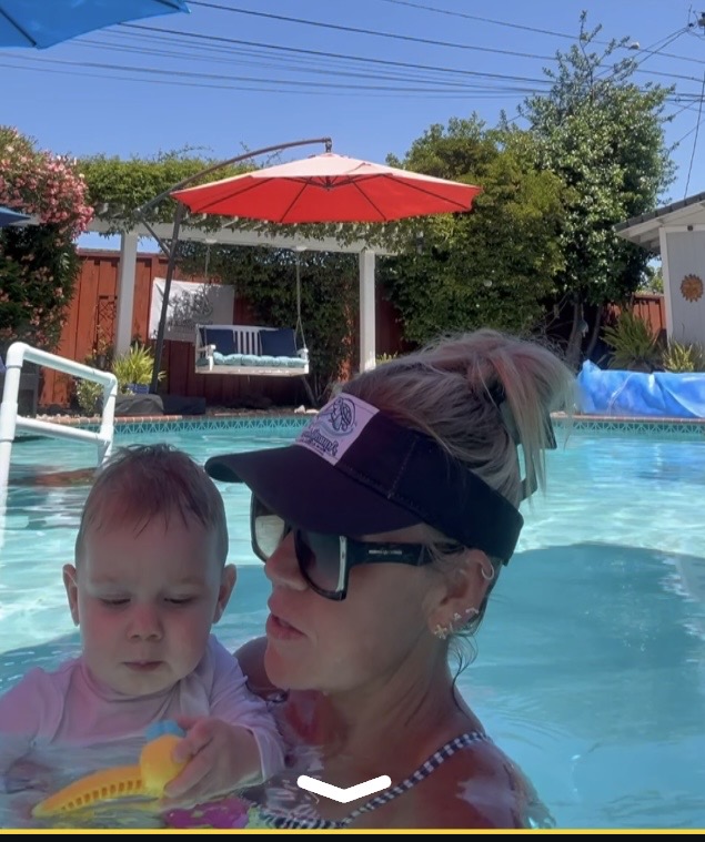 Woman and child in swimming pool, woman wearing visor and glasses, child holding a toy, poolside umbrella and trees in background.
