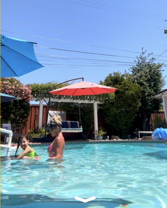 People in a swimming pool with umbrellas and trees in the background on a sunny day.