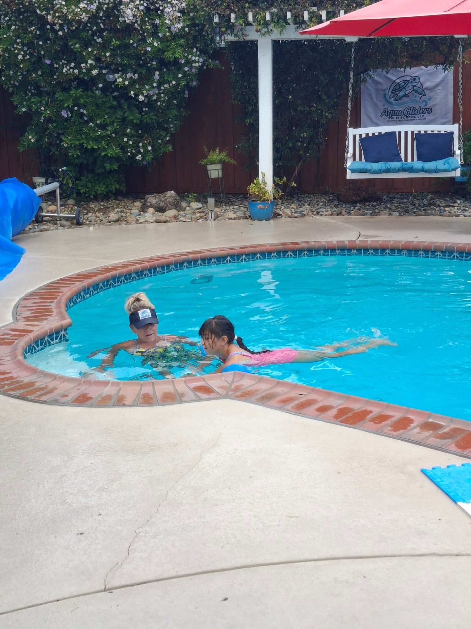 Two children swimming in a backyard pool, one wearing a swim cap, surrounded by a patio and garden with a red umbrella.