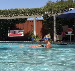 Person with sunglasses swimming in a pool with a dog, backyard with furniture and greenery in background.