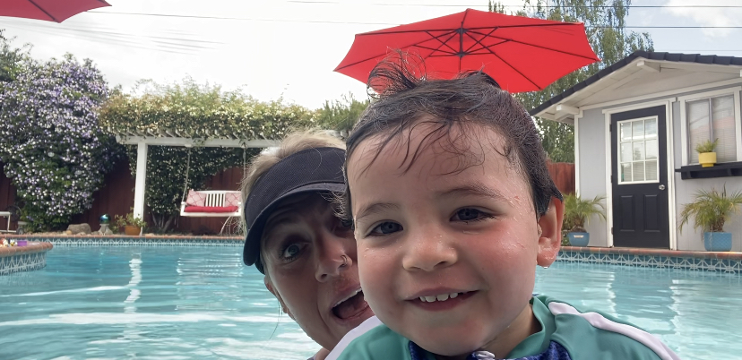Two children in a swimming pool, one smiling, with a red umbrella overhead, outdoor setting with trees and a building.