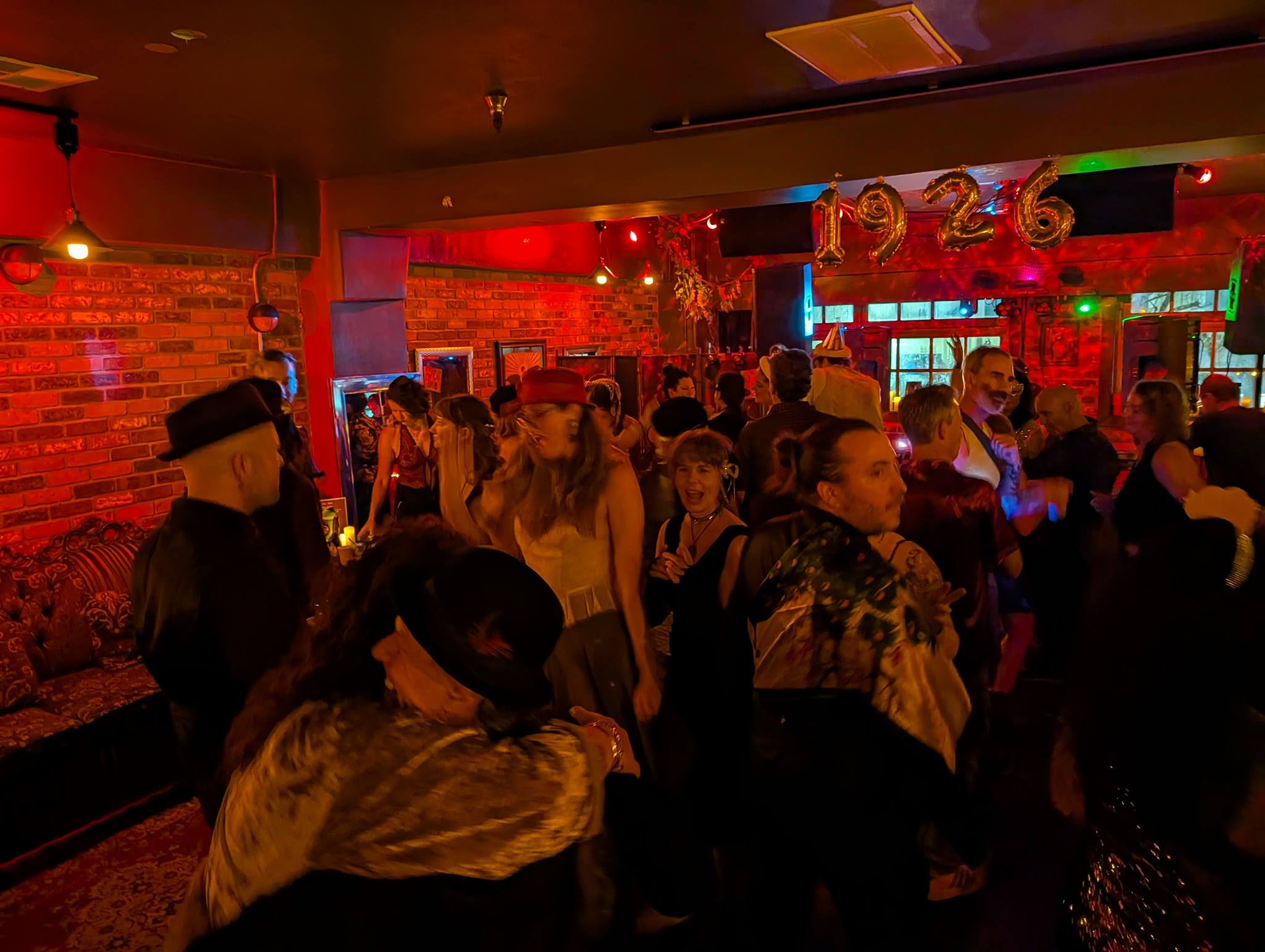 People dancing and socializing in a dimly lit bar with red brick walls and colorful lights.