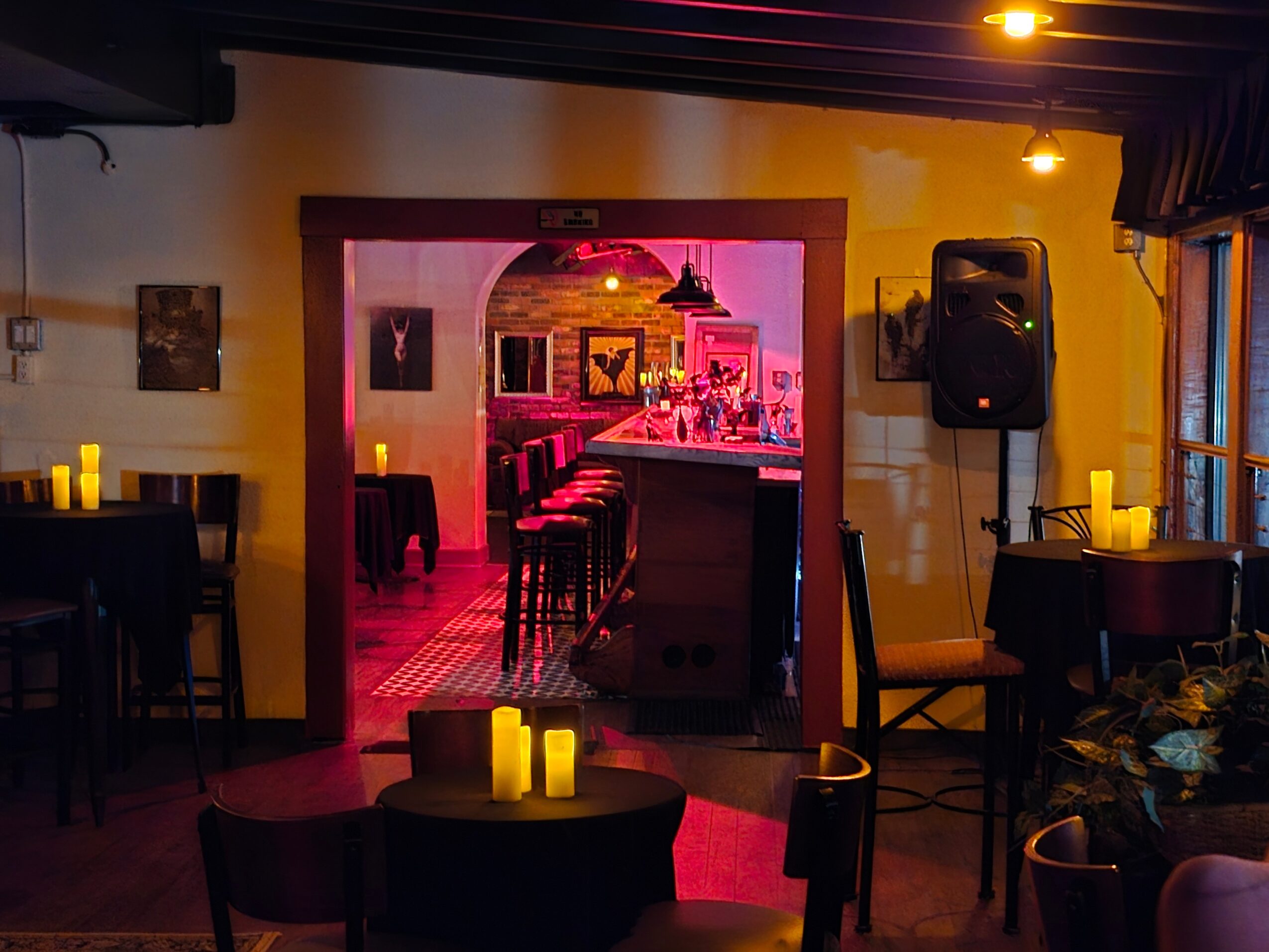 Interior of a bar or restaurant with tables, chairs, and candles, viewed through an archway with pink lighting in the background.