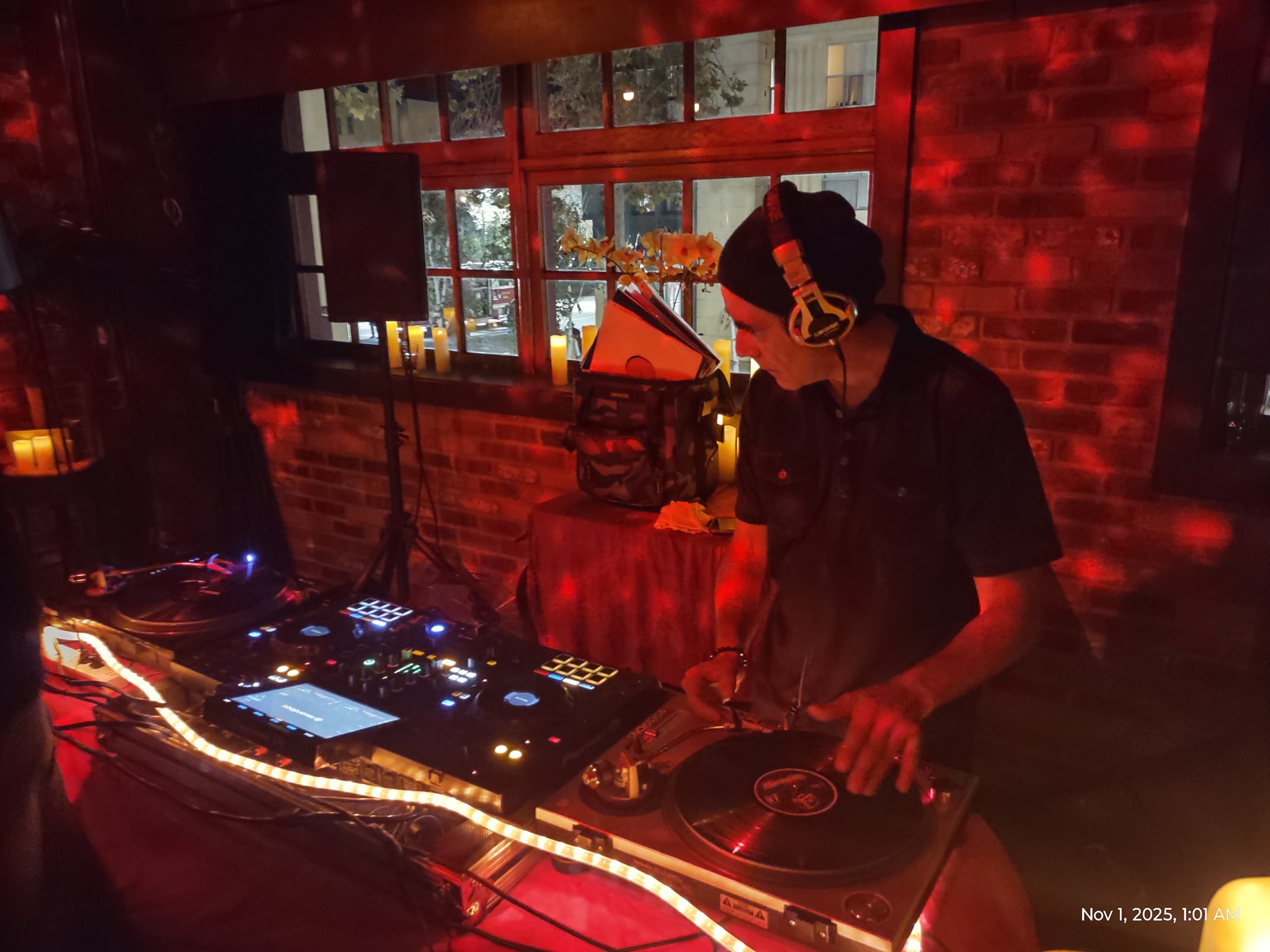 DJ wearing headphones operates turntables and equipment in a dimly lit venue with brick walls and windows.