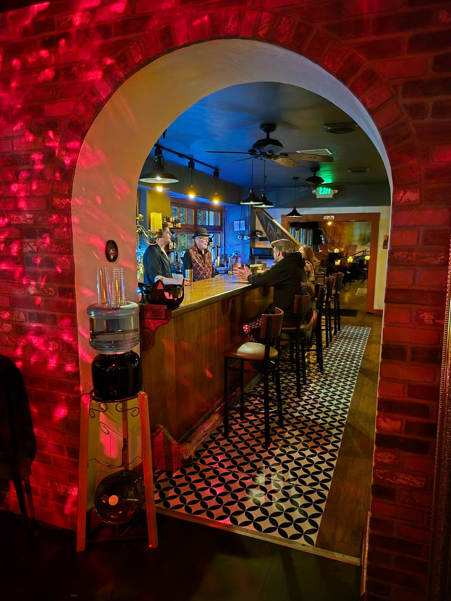 Bar with people, illuminated shelves, patterned floor, and ceiling fans, viewed through an arched doorway.