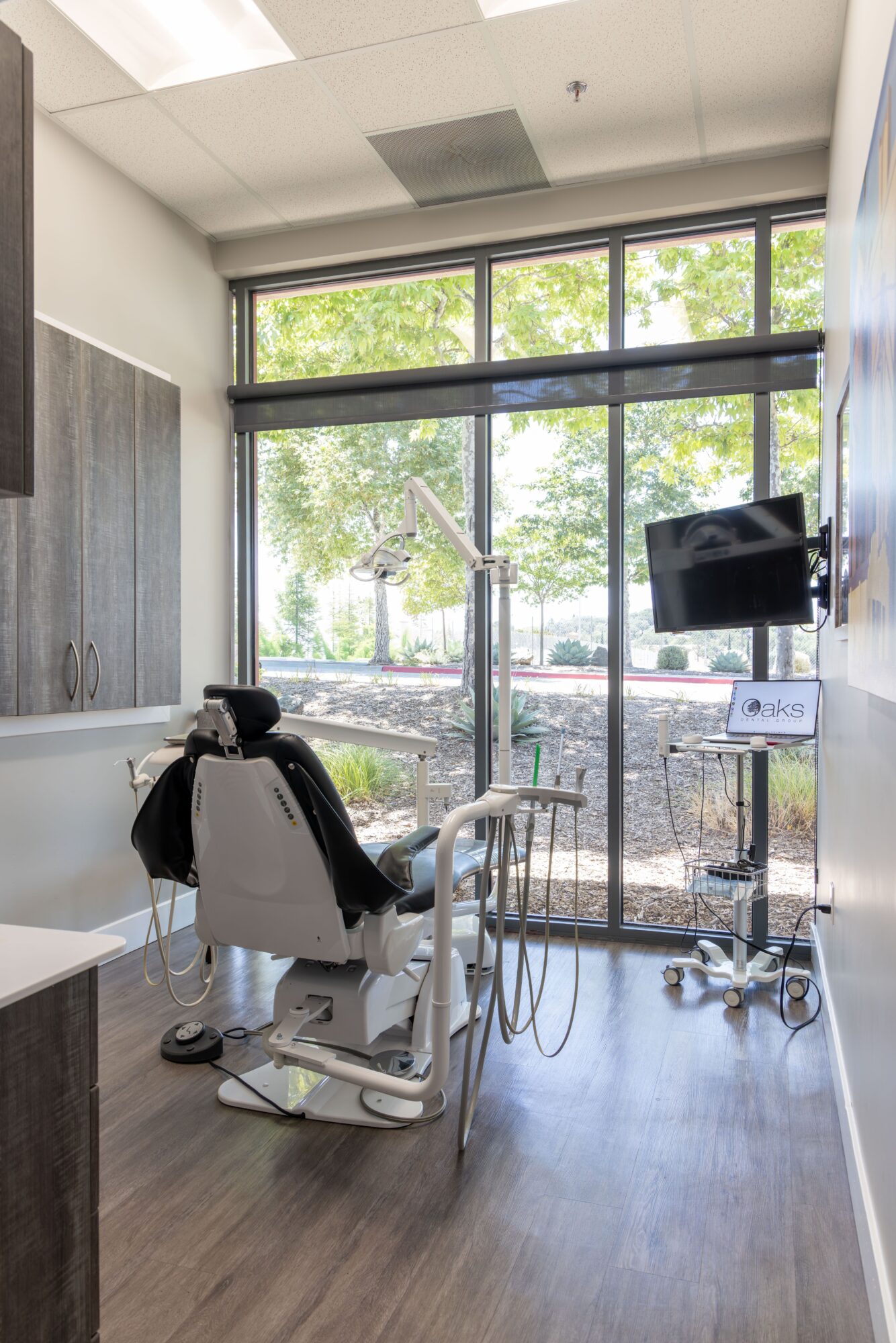 Dental chair in a treatment room with large window, trees outside, and medical equipment nearby.
