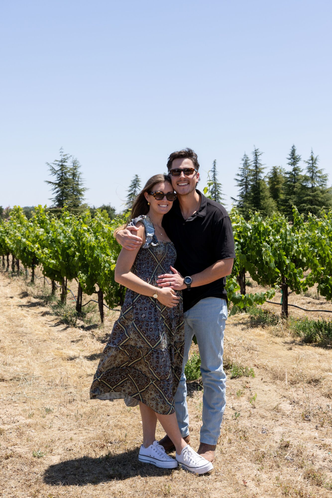 A smiling couple stands in a vineyard with green vines and trees in the background, under a clear blue sky.