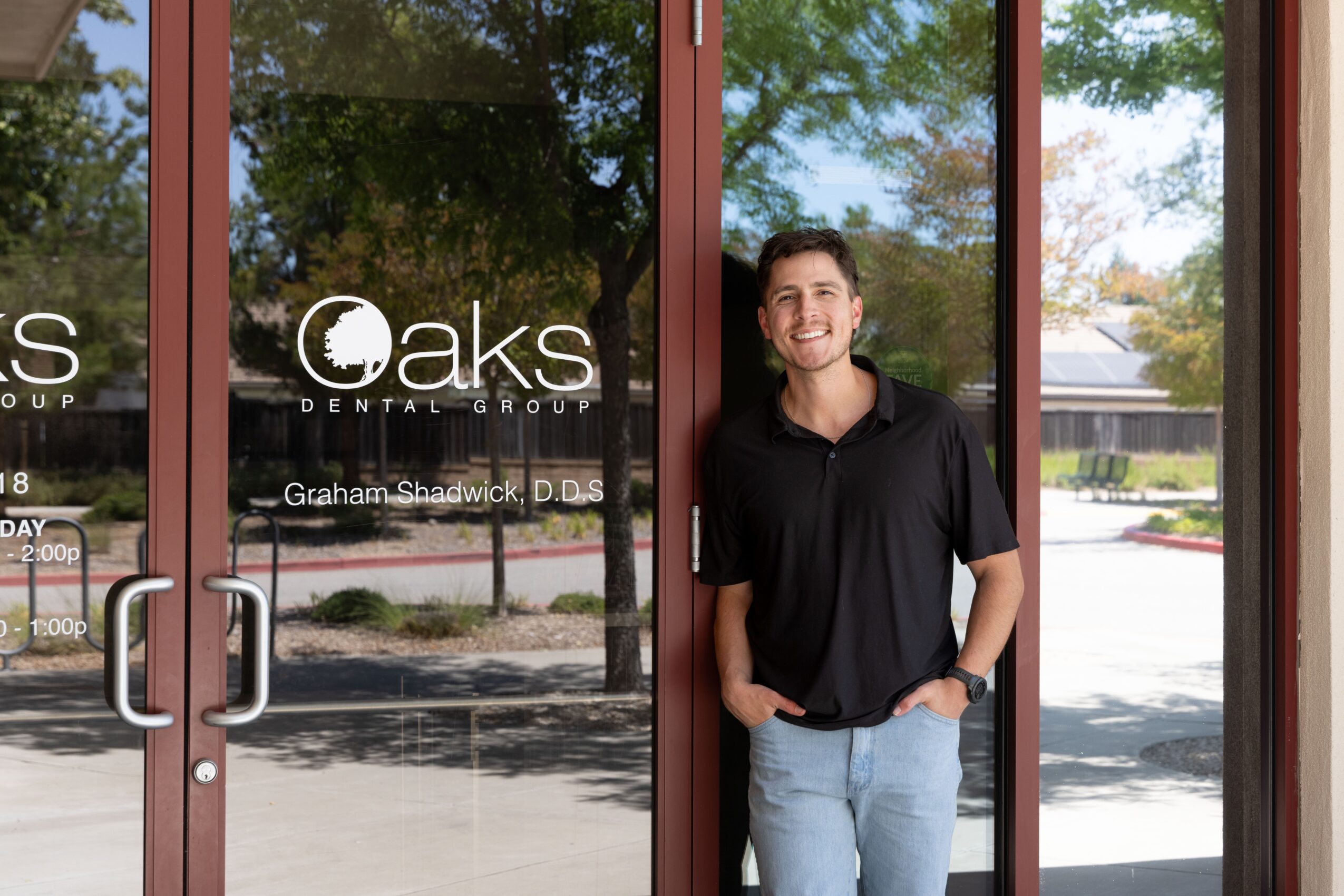 Young man standing outside a dental office, smiling, with hands in pockets, in front of glass doors with the Oaks Dental Group logo.