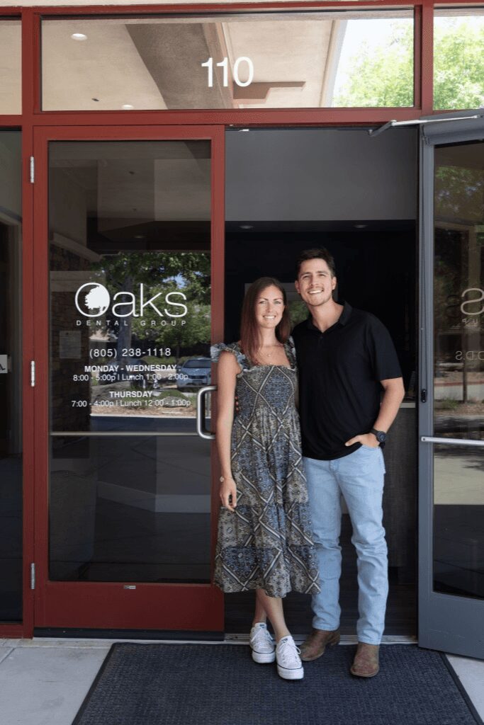 Two people stand outside a glass door with the number 110 above, smiling, with trees and reflections visible in the glass.