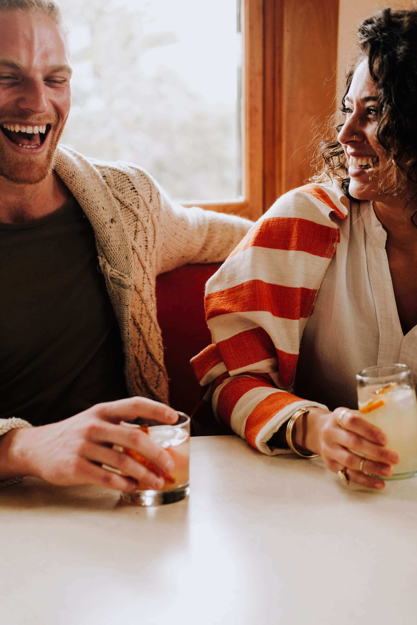 Two people sitting at a table, smiling and holding drinks, in a cozy indoor setting with a window in the background.