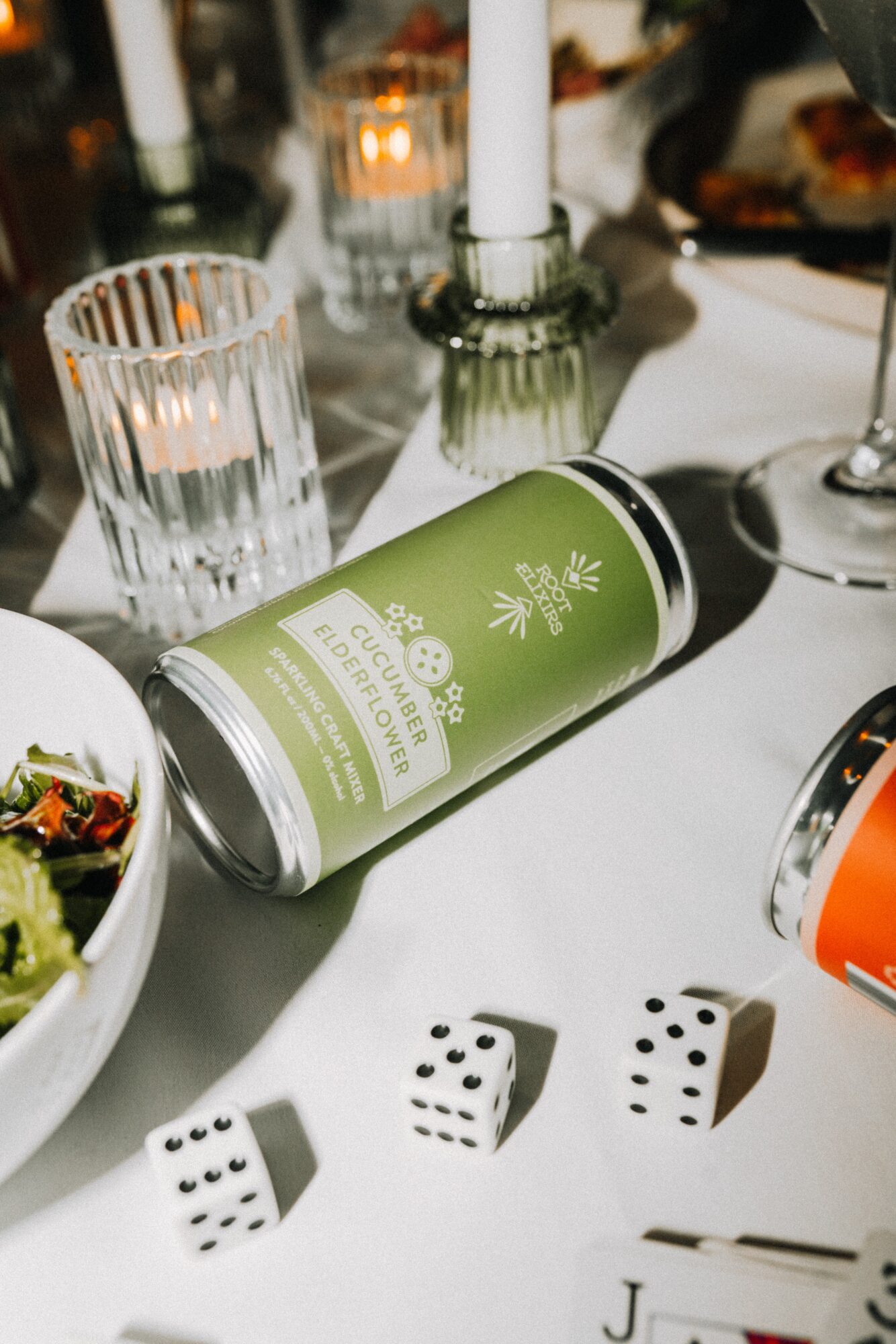 Green bottle lying on its side on a table with glasses, candles, and food, viewed from an angle.
