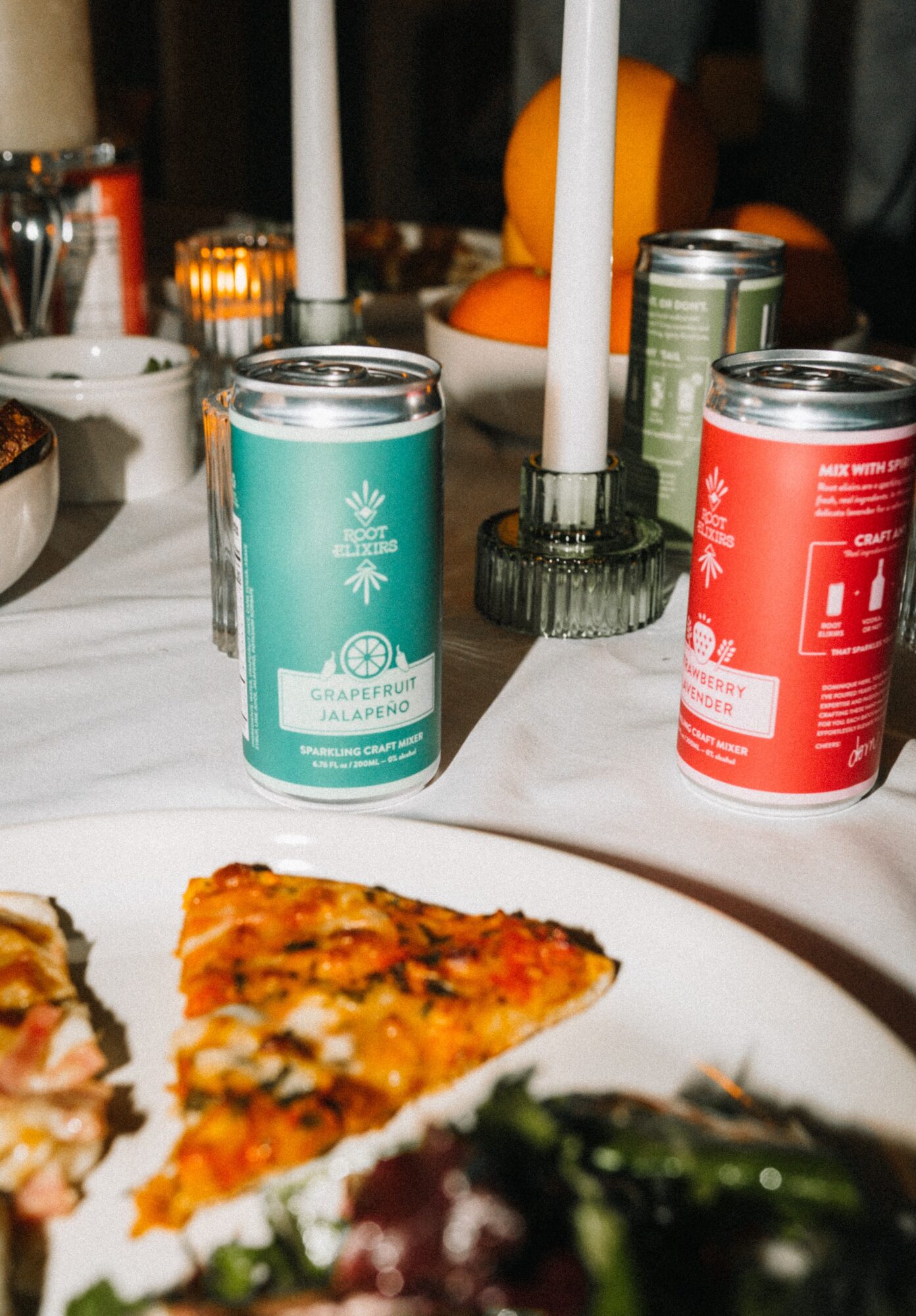 Pizza slices on a white plate with drinks, candles, and oranges in the background.