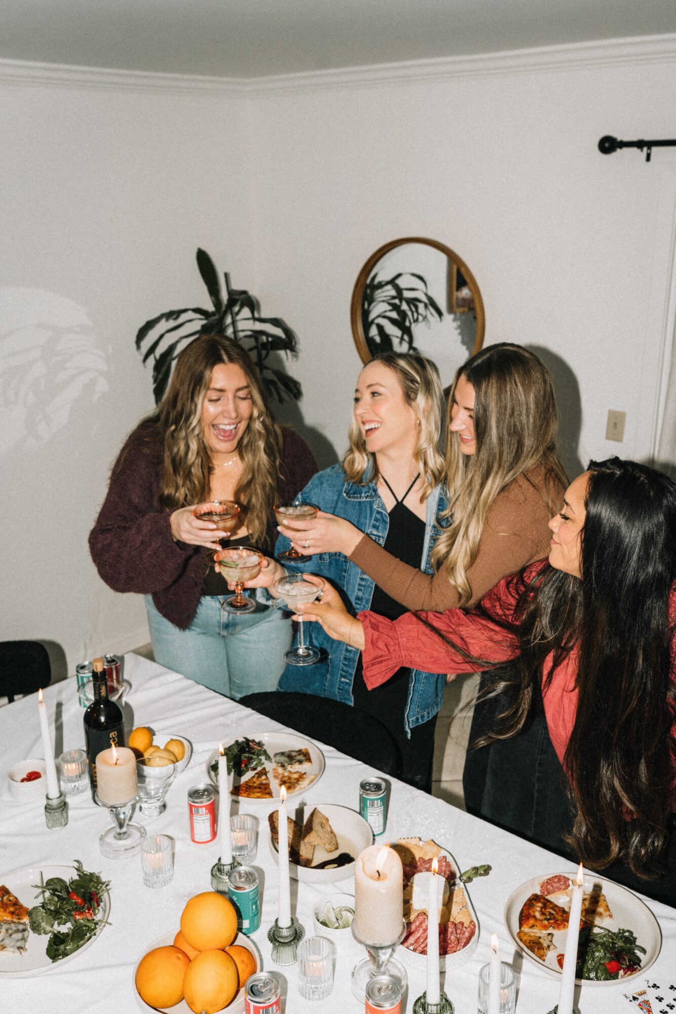 Four women gather around a table, smiling and toasting with drinks in a room with a plant and mirror.