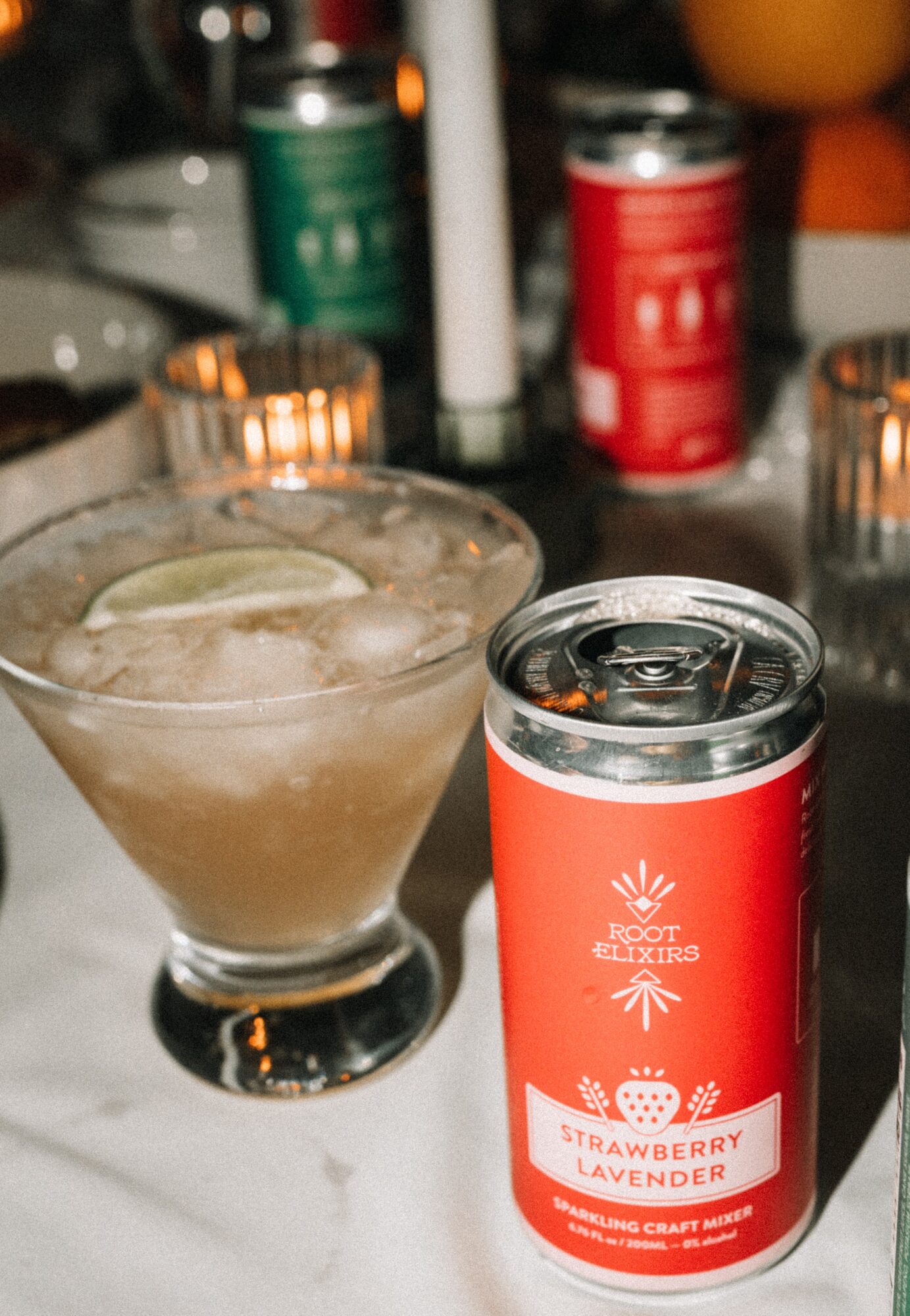 A can of strawberry lavender sparkling craft water next to a cocktail glass with ice and lime, on a table with candles.