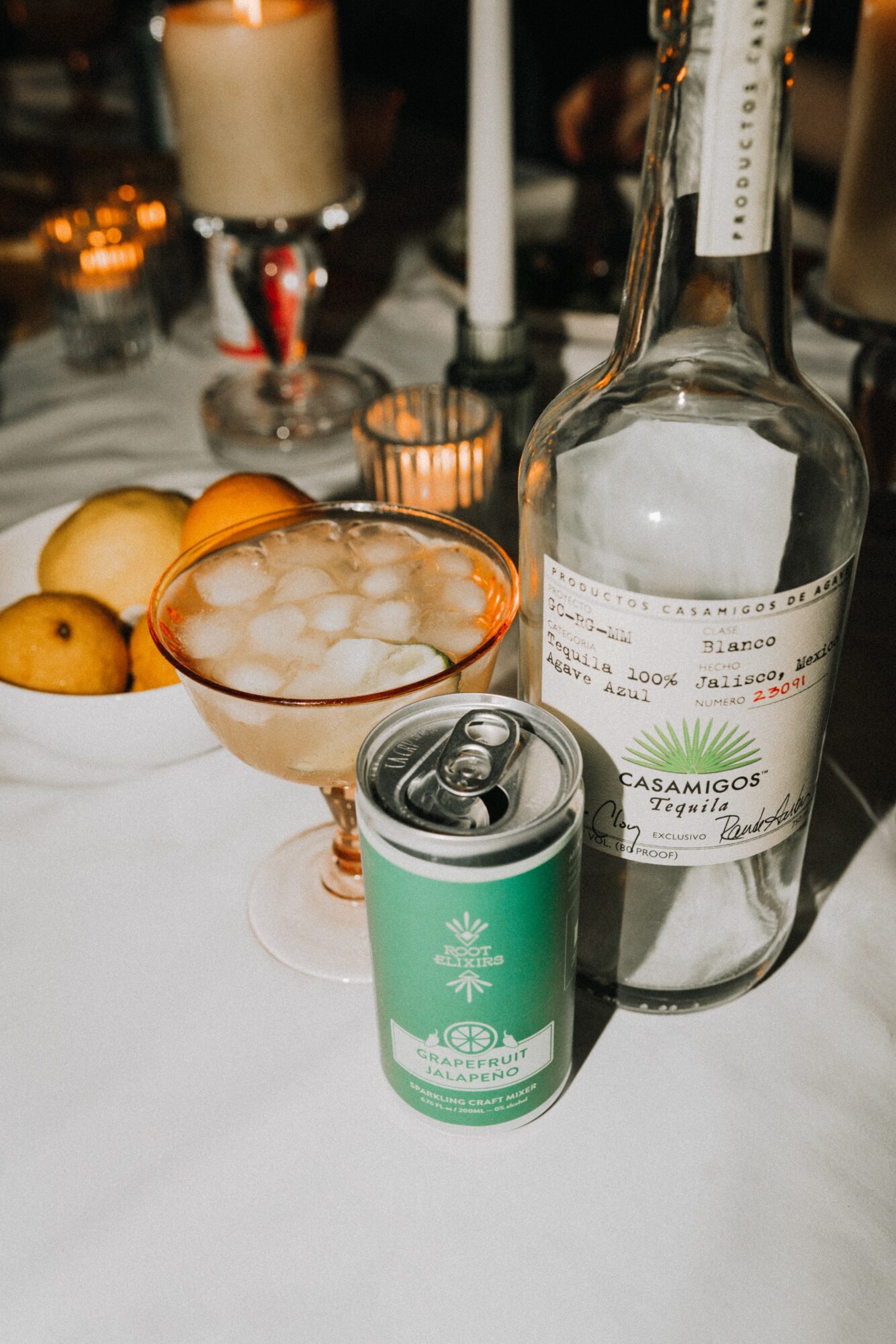 A table with a bottle, a can, a glass bowl with ice, and candles in the background.