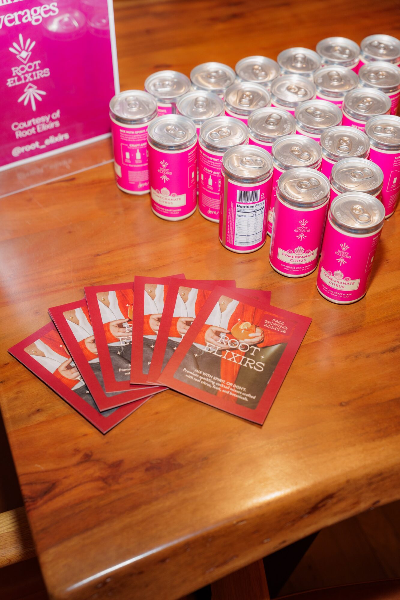 Multiple pink and silver cans arranged on a wooden table with promotional cards in front, and a pink box in the background.