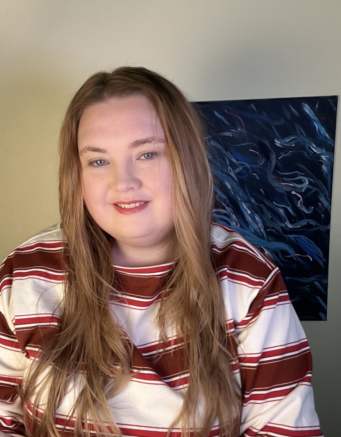 Young woman with long, wavy hair smiling, wearing a striped shirt, standing indoors with a TV in the background.