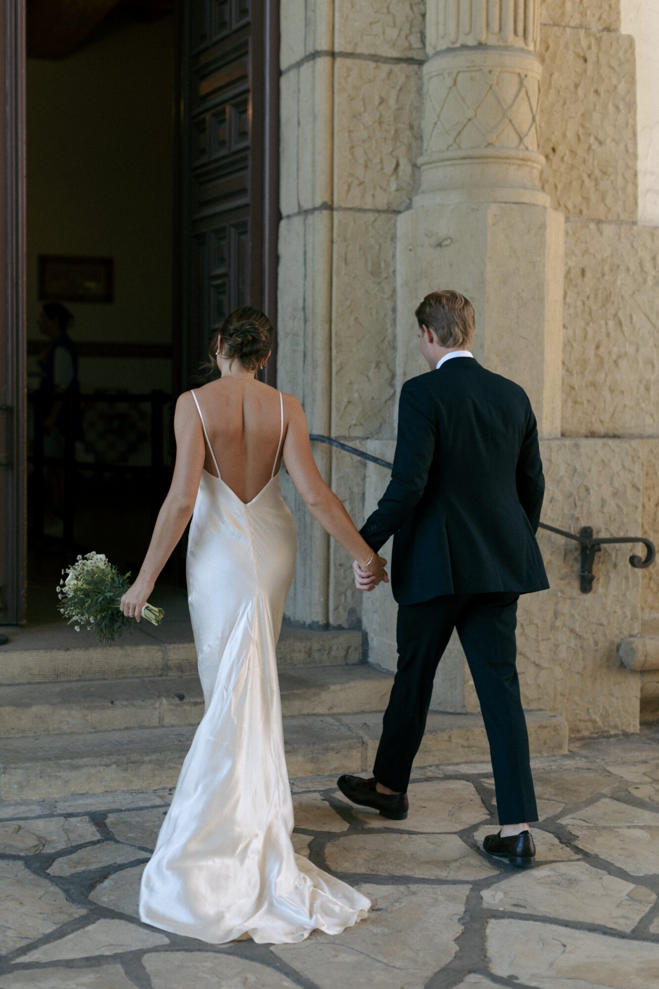 A bride and groom holding hands, walking out of a building with stone architecture, bride in a white gown, groom in a suit.