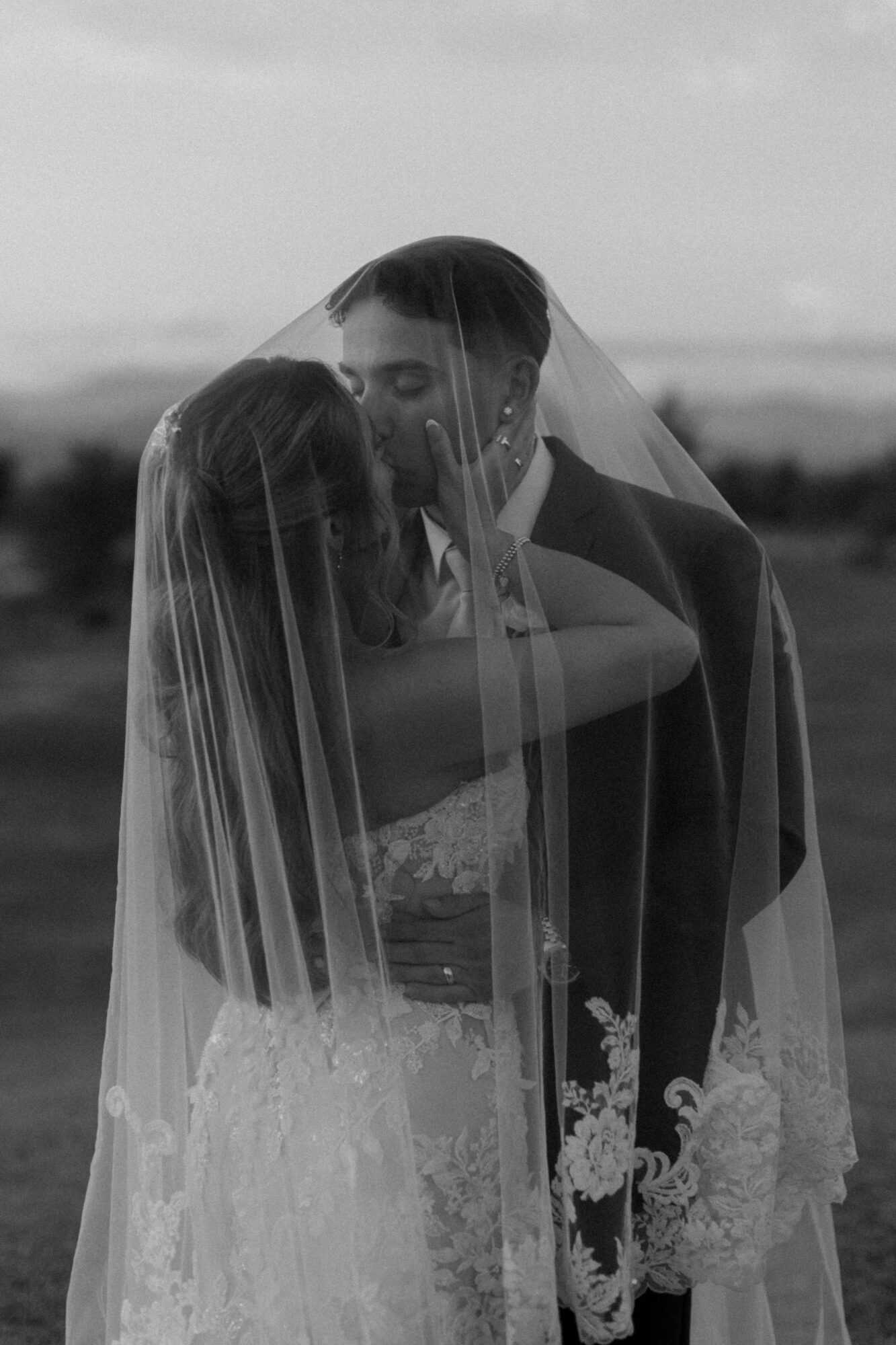 A bride and groom kiss under a veil outdoors, with a blurred landscape in the background.