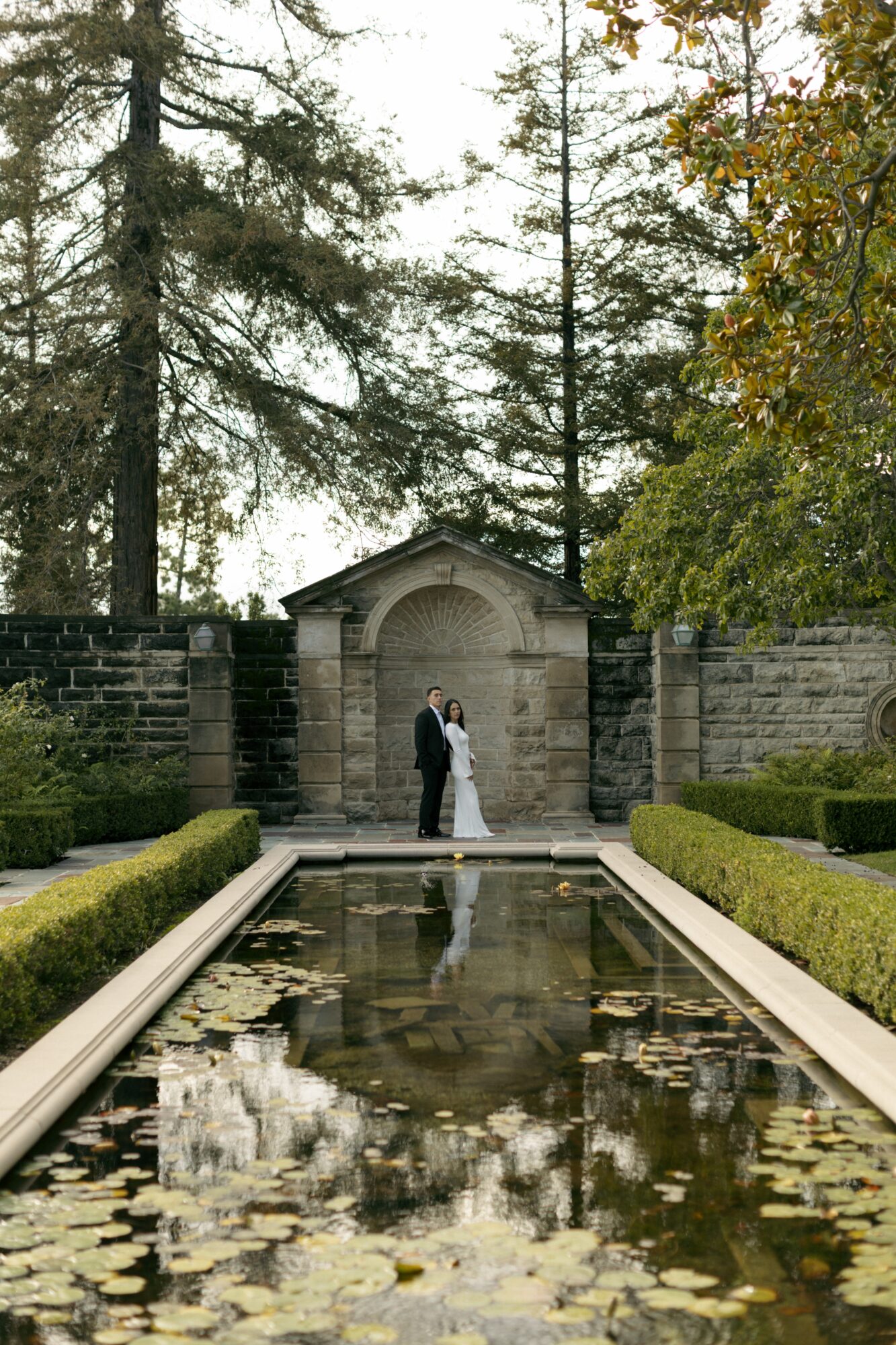 Couple standing in front of a stone archway, surrounded by trees and a reflecting pool with lily pads.