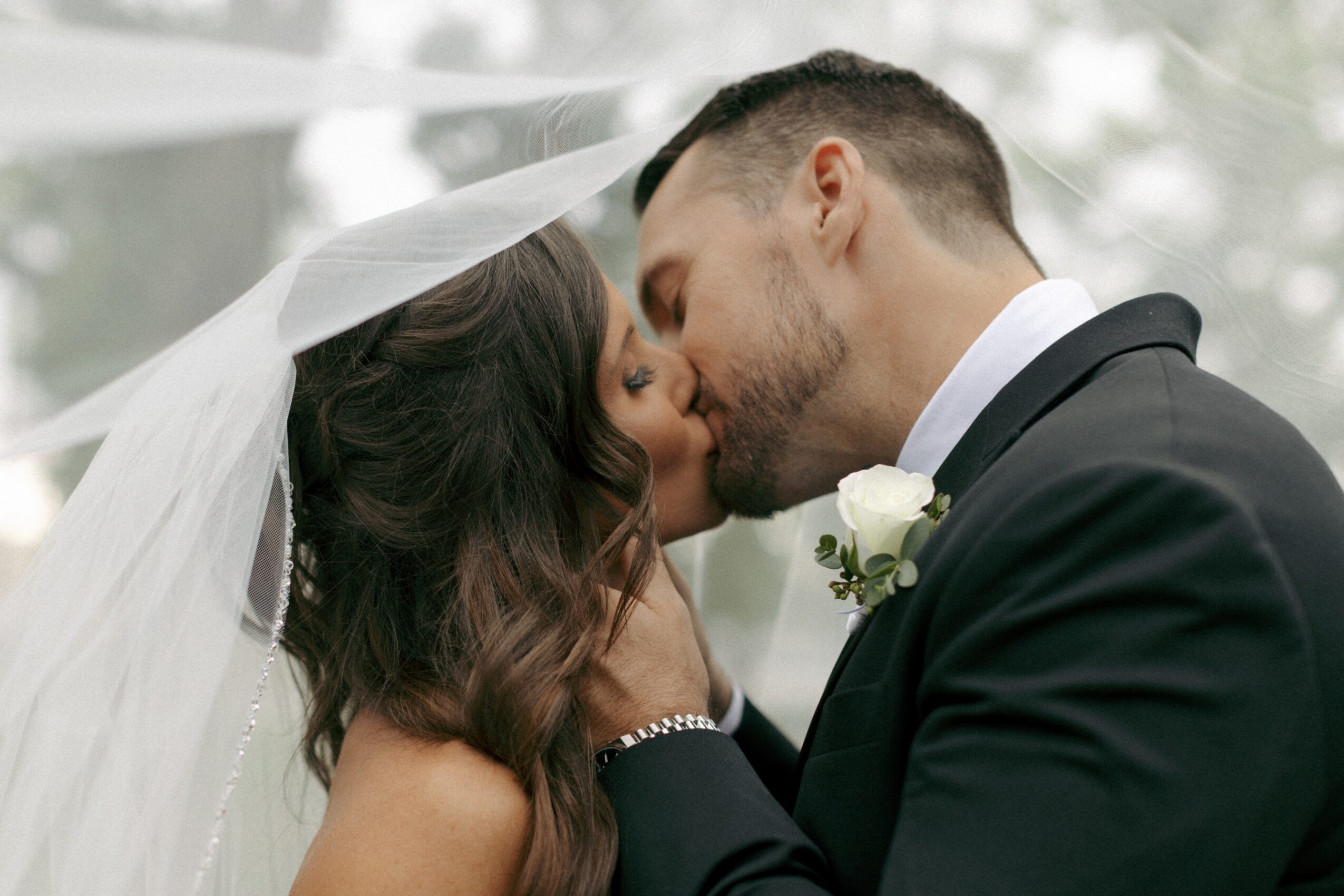A bride and groom kiss under a veil during their wedding ceremony outdoors, with trees in the background.
