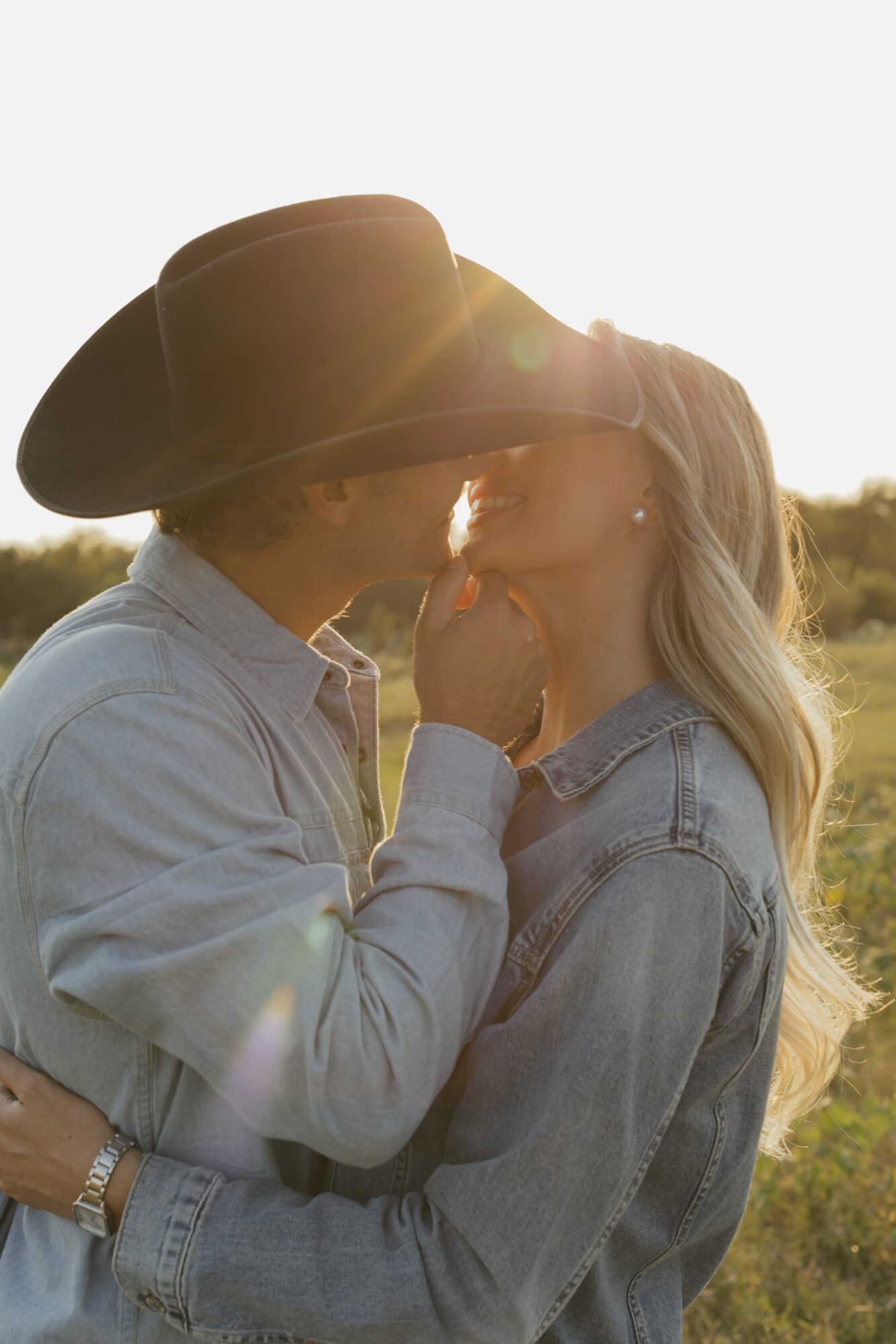 Two people share a kiss outdoors during sunset, one wearing a cowboy hat and denim jacket, the other with long hair.