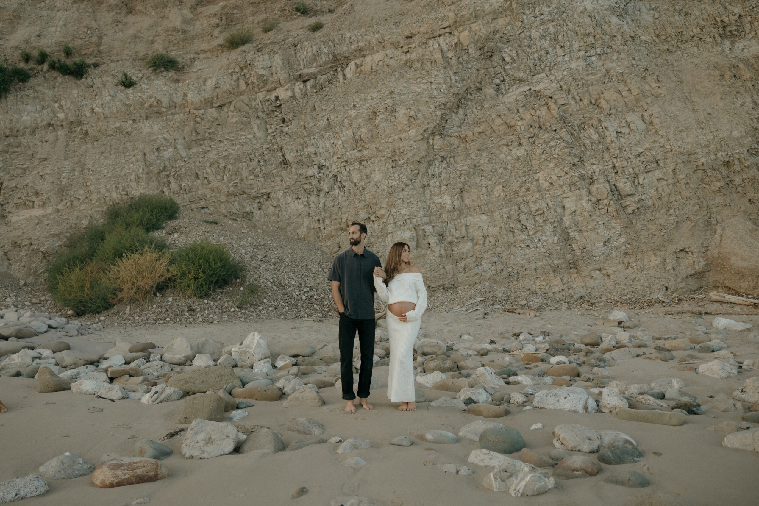 Two people stand on a rocky beach with a large cliff in the background, facing each other and holding hands.