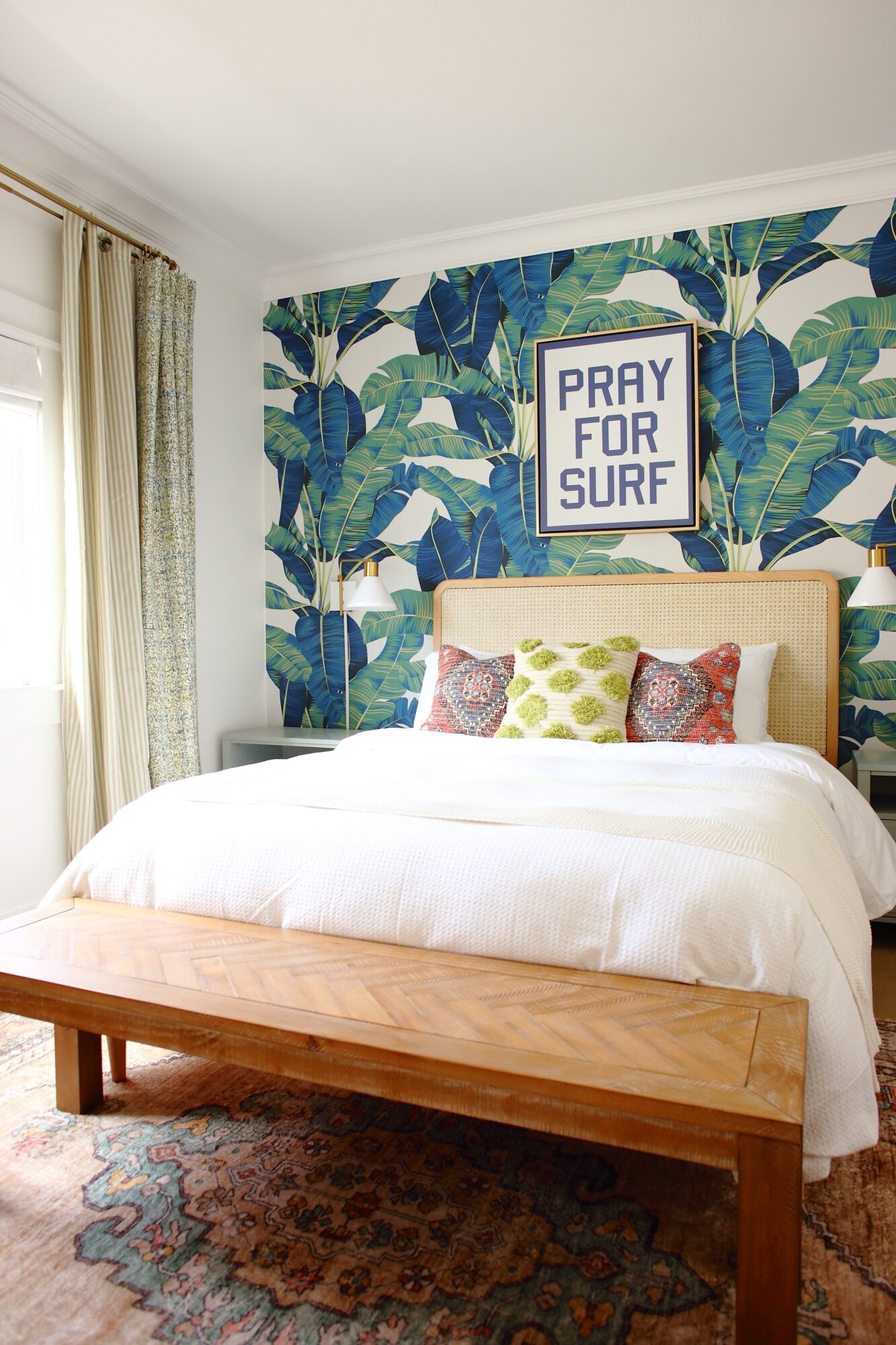 Bedroom with a tropical leaf wallpaper, a framed sign reading 'Pray for Surf,' a bed with pillows, and a wooden bench at the foot.