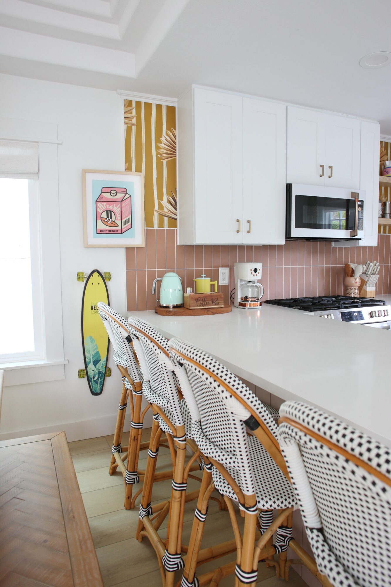 Kitchen with white cabinets, pink backsplash, and a white countertop with bar stools, a yellow surfboard, and kitchen appliances.