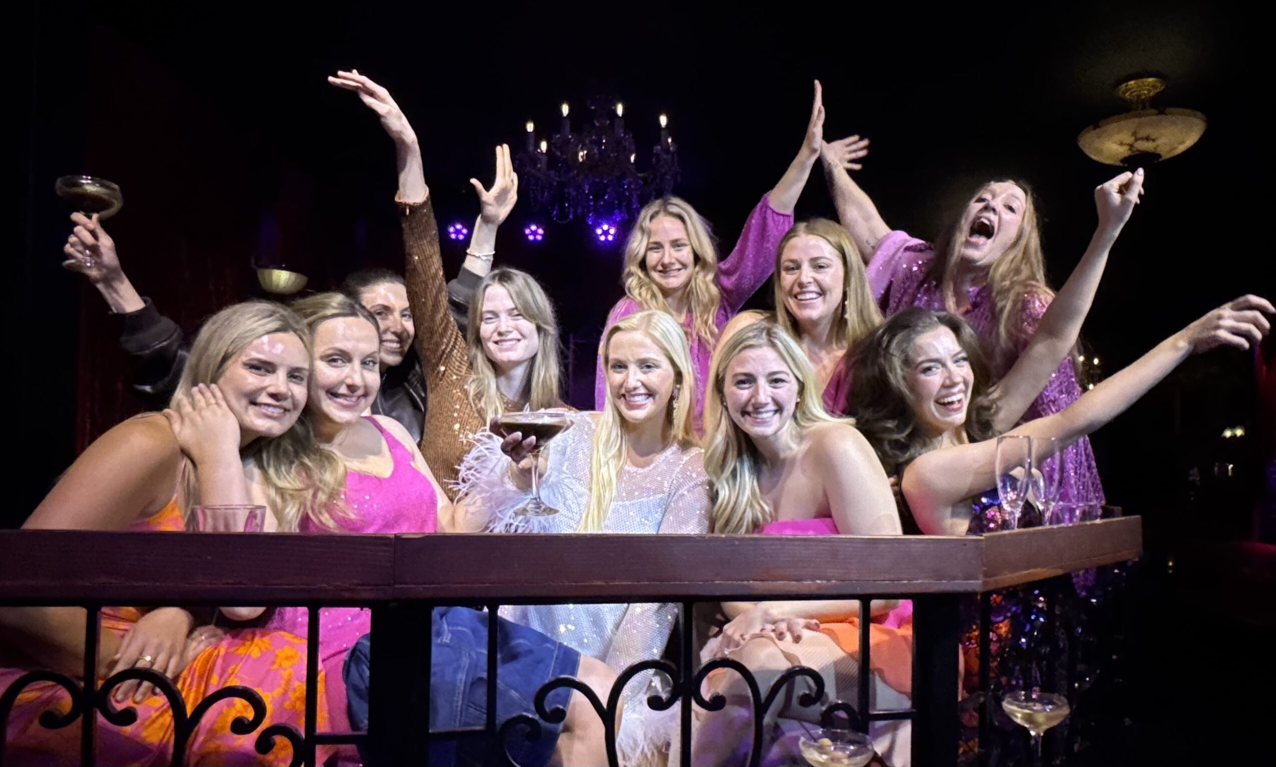 Group of women celebrating on a balcony, smiling and raising their hands, with purple lighting in the background.