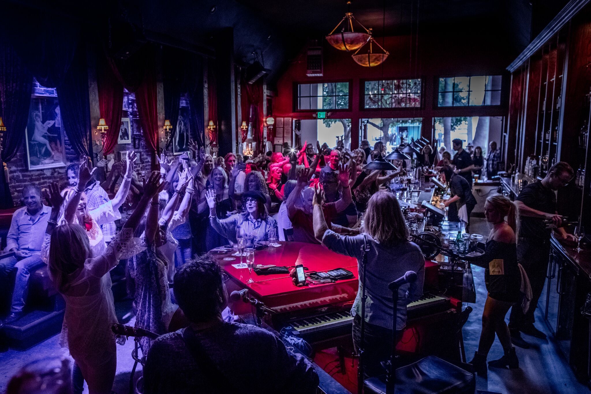 Crowded indoor party with people dancing, socializing, and enjoying drinks, illuminated by colorful lighting and large windows in the background.
