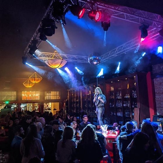 Performer on stage with colorful lights, audience watching in a lively indoor venue, disco ball overhead, people seated and standing.