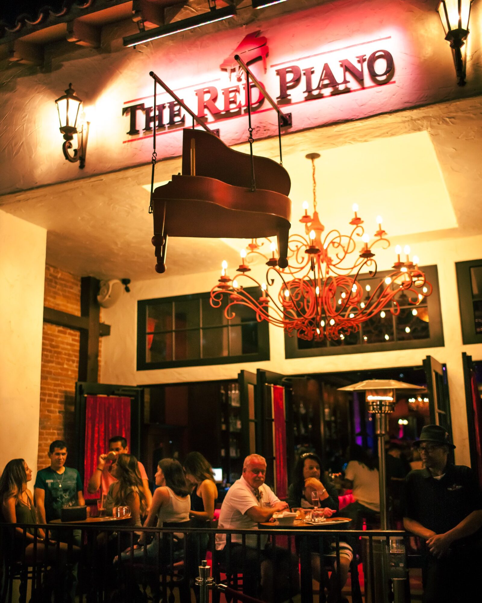 Interior of a restaurant with a grand piano hanging from the ceiling, chandelier, and people dining at tables.