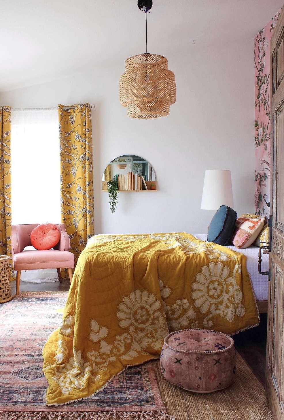 Bedroom with yellow floral bedspread, pink chair, window with yellow curtains, and pink pouf, decorated with wall art and a lamp.