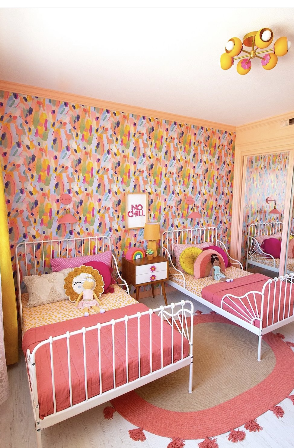 Two white metal beds with pink bedding and stuffed animals, colorful wall, and a window with curtains in a children's room.